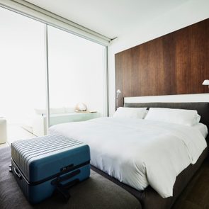 Suitcase sits on a bench beside a bed in a modern, bright bedroom with a wooden headboard and sliding glass doors.