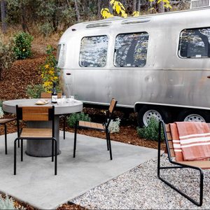 Table surrounded by chairs holds wine and bread, situated outdoors near a silver trailer with a forested background.