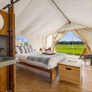 Bed with pillows and blanket inside a glamping tent, set in a grassy field with wooden chairs outside.