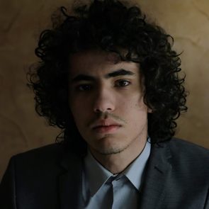 A person with curly hair stares intently, wearing a suit, against a muted brown background.