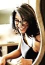 A smiling person with glasses leans forward, indoors, near a desk with blurred items, creating a warm and inviting atmosphere.