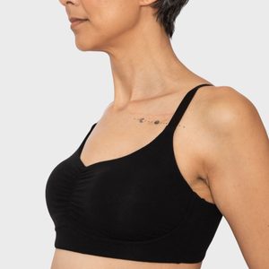 Woman wearing a black bra, standing sideways, with a small solar system tattoo on her upper chest, against a plain background.
