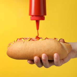 A hand holds a hot dog being topped with ketchup and mustard. The background is a bright yellow.