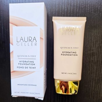 A tube of foundation stands beside its box on a dark surface, labeled "Laura Geller Quench-N-Tint Hydrating Foundation" with weightless coverage.