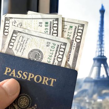 Hand holds a passport with hundred-dollar bills, against a window view of the Eiffel Tower in the background.