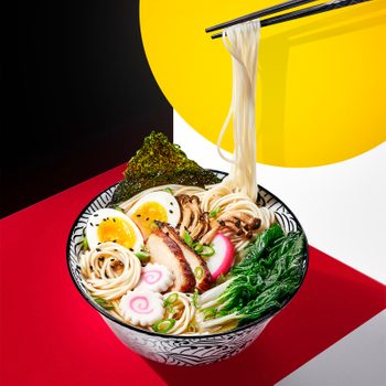 Bowl of ramen contains noodles, egg, seaweed, pork, and greens; chopsticks lift noodles, set against a bold black, red, and yellow geometric background.