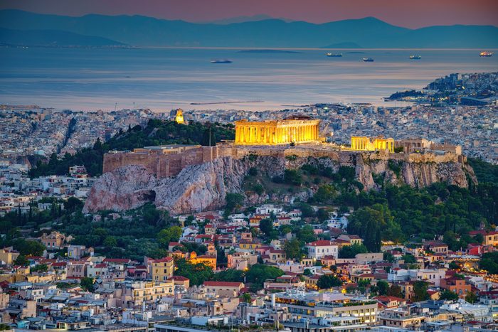 The illuminated Parthenon stands atop the Acropolis, overlooking Athens at dusk with distant mountains and sea.