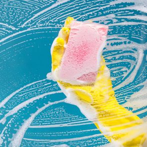 A yellow-gloved hand holds a pink sponge, cleaning a soapy, streaked glass surface.