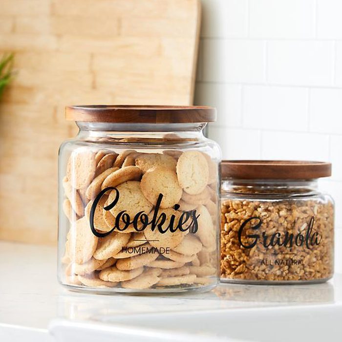 Two glass jars hold cookies and granola on a kitchen counter, with wooden lids and labeled text in a bright, tiled setting.