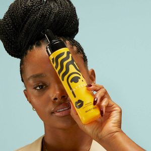 A woman holds a yellow hair product bottle, with black abstract designs, against a light blue background. Text: 