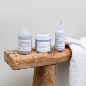 Three hair care bottles sit on a wooden bench beside a white towel, each labeled with 
