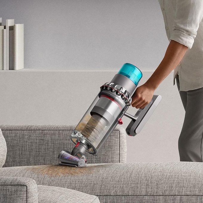 A hand-held vacuum cleans a gray sofa, picking up dust and dirt, in a modern living room setting with a minimalist bookshelf.