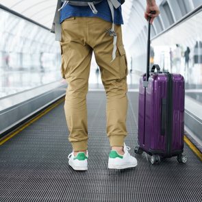 Traveler pulling purple suitcase on moving walkway in a modern airport terminal, wearing tan pants and white sneakers with green accents.