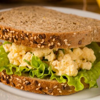 A sandwich sits on a plate, featuring multigrain bread, scrambled eggs, and fresh lettuce, placed indoors.