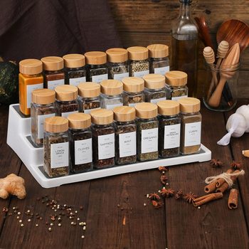 Spice jars with wooden lids sit organized on a tiered tray, surrounded by kitchen utensils and ingredients on a wooden surface.