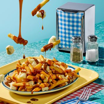 Poutine with gravy drips onto fries and cheese curds on a yellow tray. Salt, pepper, and napkin holder rest on a turquoise table.