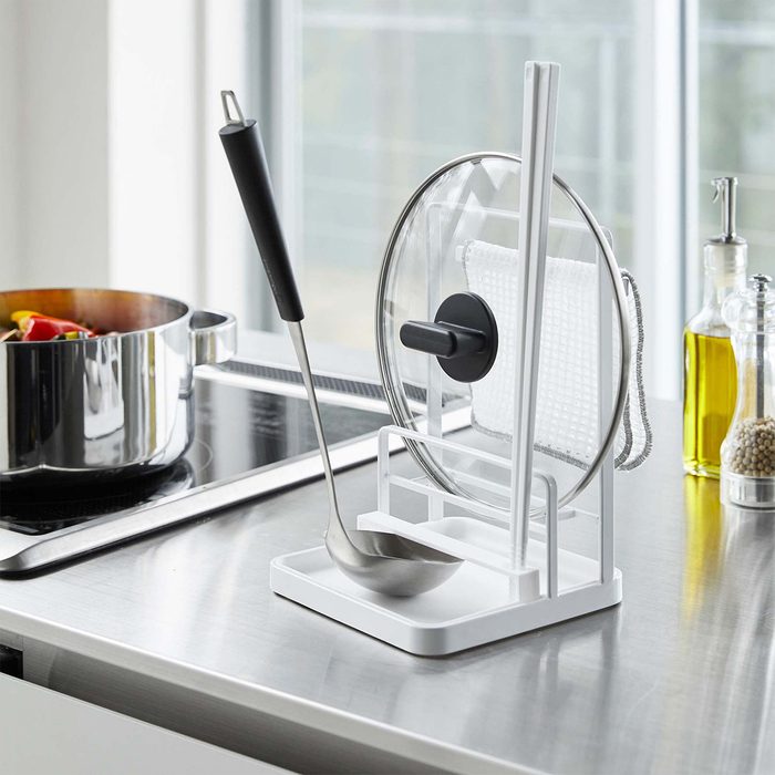 Utensil holder secures ladle and pot lid, standing on a kitchen countertop. Nearby, a pot simmers on the stove and bottles with oil sit behind.