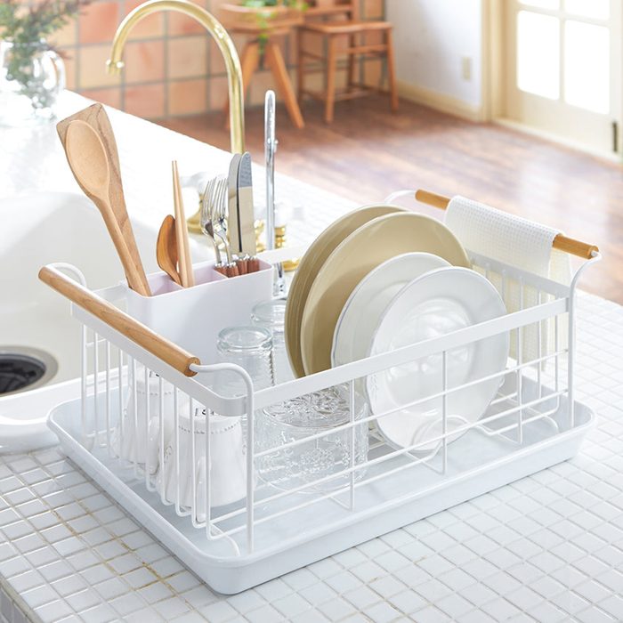 Dish rack holds plates, utensils, and cups on a white tiled countertop in a bright kitchen setting.
