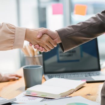 Two people shake hands over a wooden desk with papers, a laptop, and a cup in an office setting.