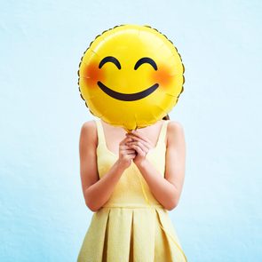 Balloon held covering face, with yellow smiley design, person wearing matching yellow dress, against a light blue background.