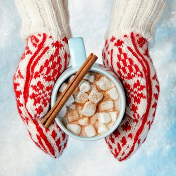 Mug filled with hot chocolate and marshmallows, held by gloved hands, decorated with warm red patterns, resting on a snowy background.