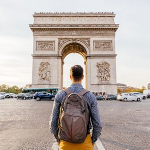 Backpack-wearing person stands observing the Arc de Triomphe as cars drive past on a busy street, surrounded by autumn-colored trees.