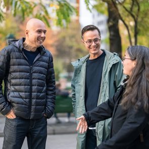 Three people conversing happily in a park, surrounded by trees and blurred foliage, with one person gesturing animatedly.