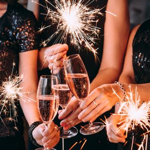 Sparklers illuminating raised champagne glasses, amidst people in glittery, festive attire, suggesting a celebratory event or party atmosphere.