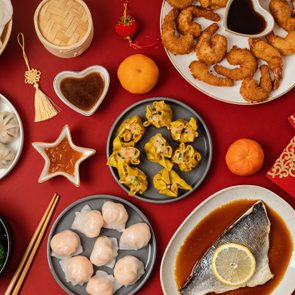 Assorted Chinese dishes arranged on a red table, featuring dumplings, shrimp, fish, vegetables, sauces, tangerines, chopsticks, and festive decorations.