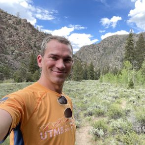 Man smiling, wearing an orange UTMB 2018 shirt, stands in a lush, mountainous area with trees and a bright blue sky overhead.
