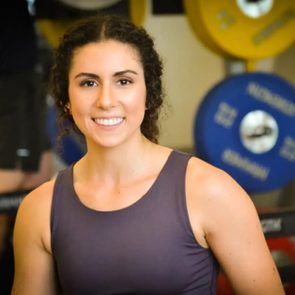 A person smiling, standing in front of weightlifting equipment in a gym setting.