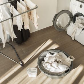 Laundry basket holds clothes, washer with open door nearby, clothes drying on a rack in sunlit laundry room.
