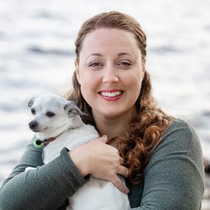 A person smiles while holding a small dog, set against a blurred water background.