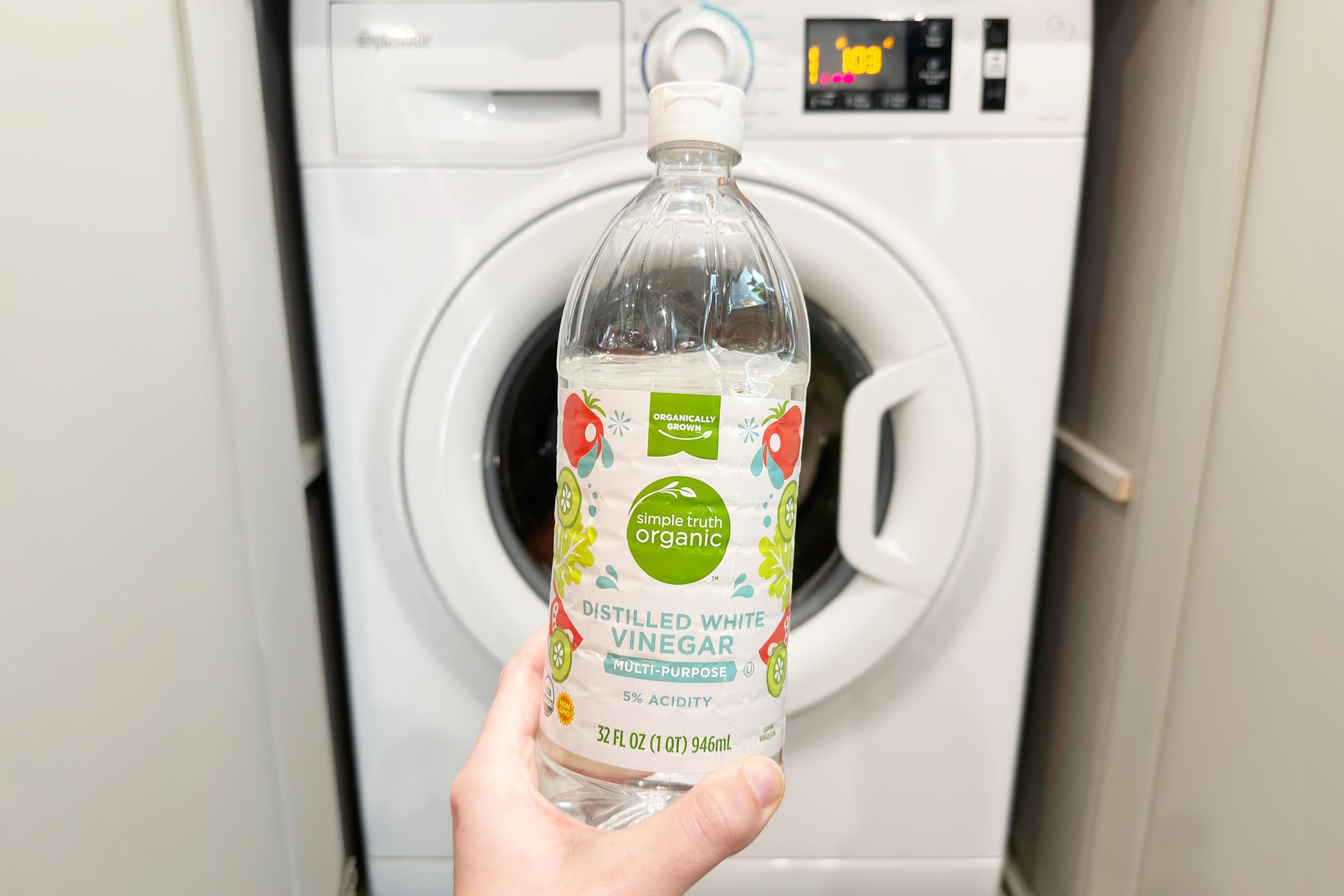 A bottle of distilled white vinegar is held in front of a washing machine in a laundry room.