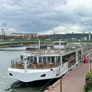River cruise ship docked, anchored by a quay in an urban riverside setting, with a bridge and cityscape under a cloudy sky.