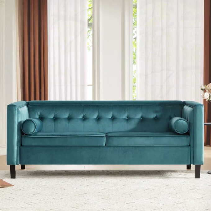 Teal velvet sofa sits on a beige rug, bordered by brown curtains and large windows showing trees outside.