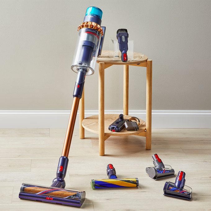 A cordless vacuum leans against a light wood table, surrounded by various interchangeable cleaning heads on a wooden floor.