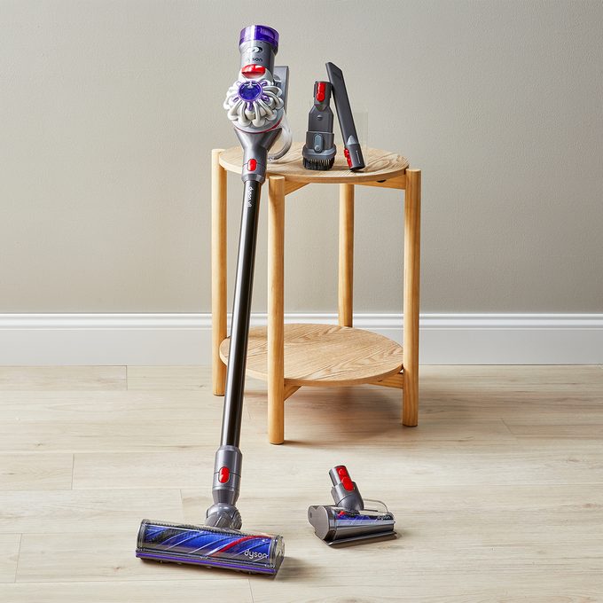 Vacuum cleaner stands with attachments arranged on a wooden table against a plain indoor wall on a wooden floor.