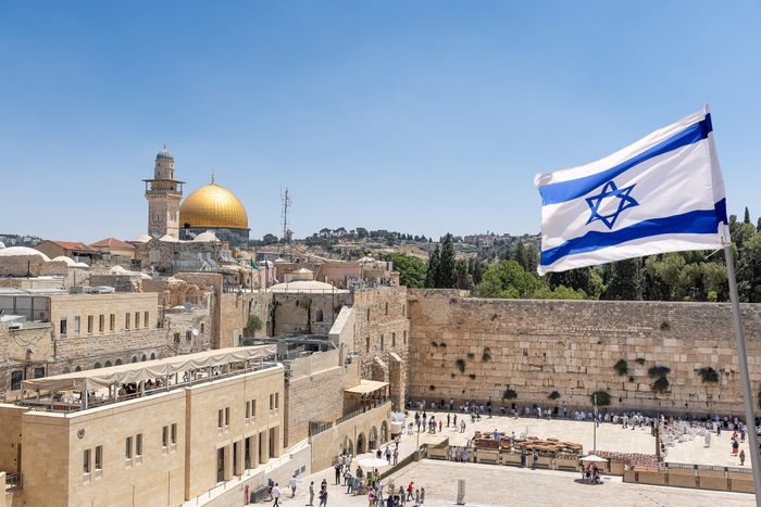 Israeli flag flutters above the Western Wall, with the golden Dome of the Rock and Jerusalem's historic architecture in the background under a clear sky.
