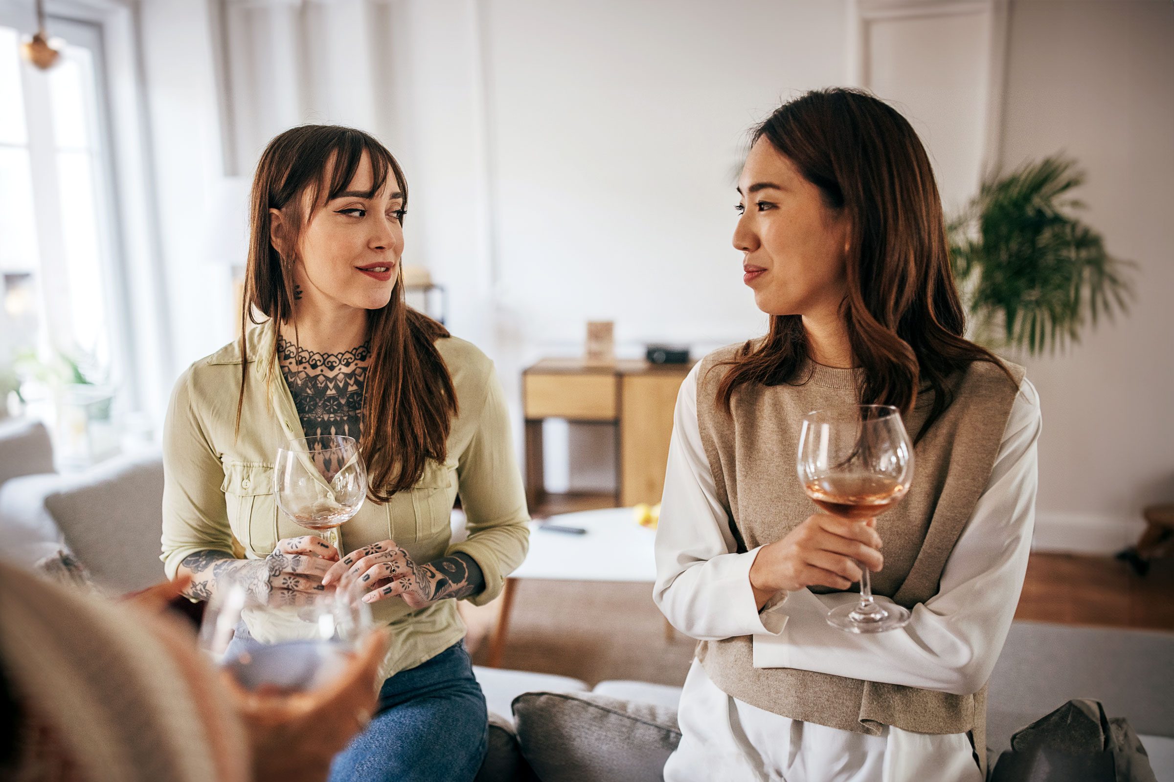 Two people are holding wine glasses, conversing casually in a modern, indoor living space with light decor and a potted plant.
