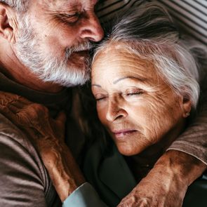Elderly couple cuddling warmly, eyes closed, resting on striped bedding.