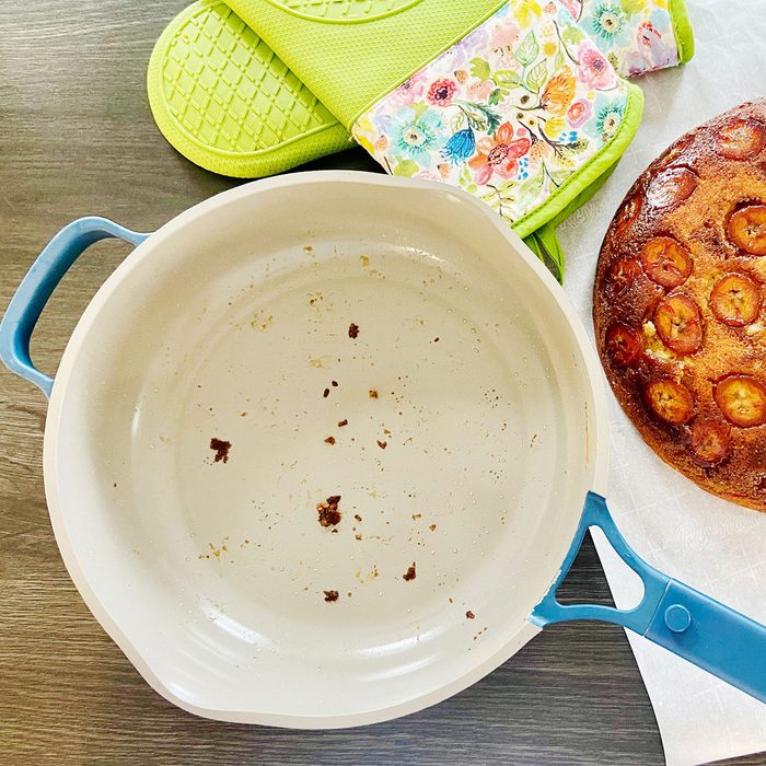 Pan with food crumbs rests on a gray surface, beside green oven mitts and banana-topped, round baked cake.