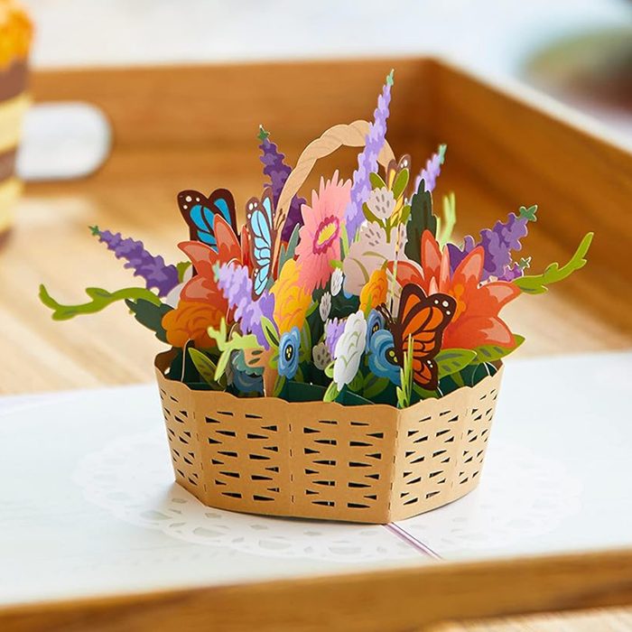 Paper flowers and butterflies emerge from a woven basket, presented on a wooden tray in a bright setting.