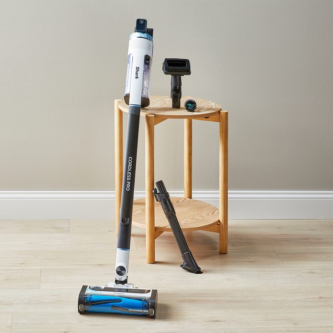 Vacuum cleaner stands upright on wooden floor; additional attachments rest on two-tier wooden cart against a light-colored wall.
