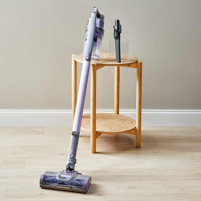 A cordless vacuum stands upright, leaning against a wooden side table, in a room with a light wooden floor and plain wall.