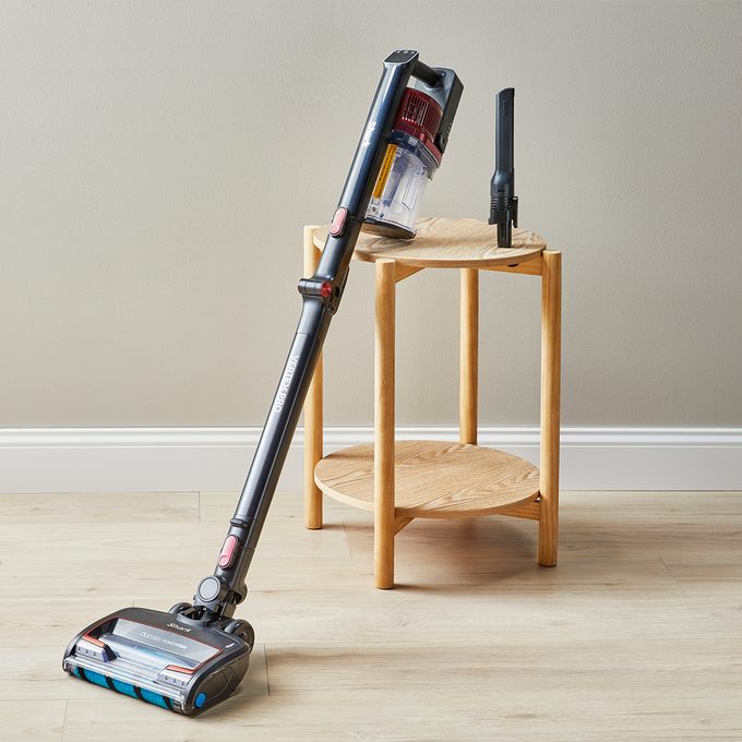 Vacuum stands upright on wooden table in brightly lit room, adjacent to beige wall and light wooden floor.