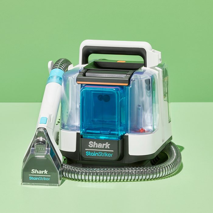Carpet cleaner rests on light green surface, featuring a hose and transparent compartments, with text 