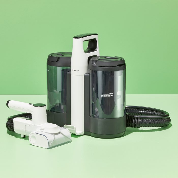 Vacuum cleaner rests on a light green surface, featuring two cylindrical elements and a detachable nozzle with a coiled hose, set against a green background.