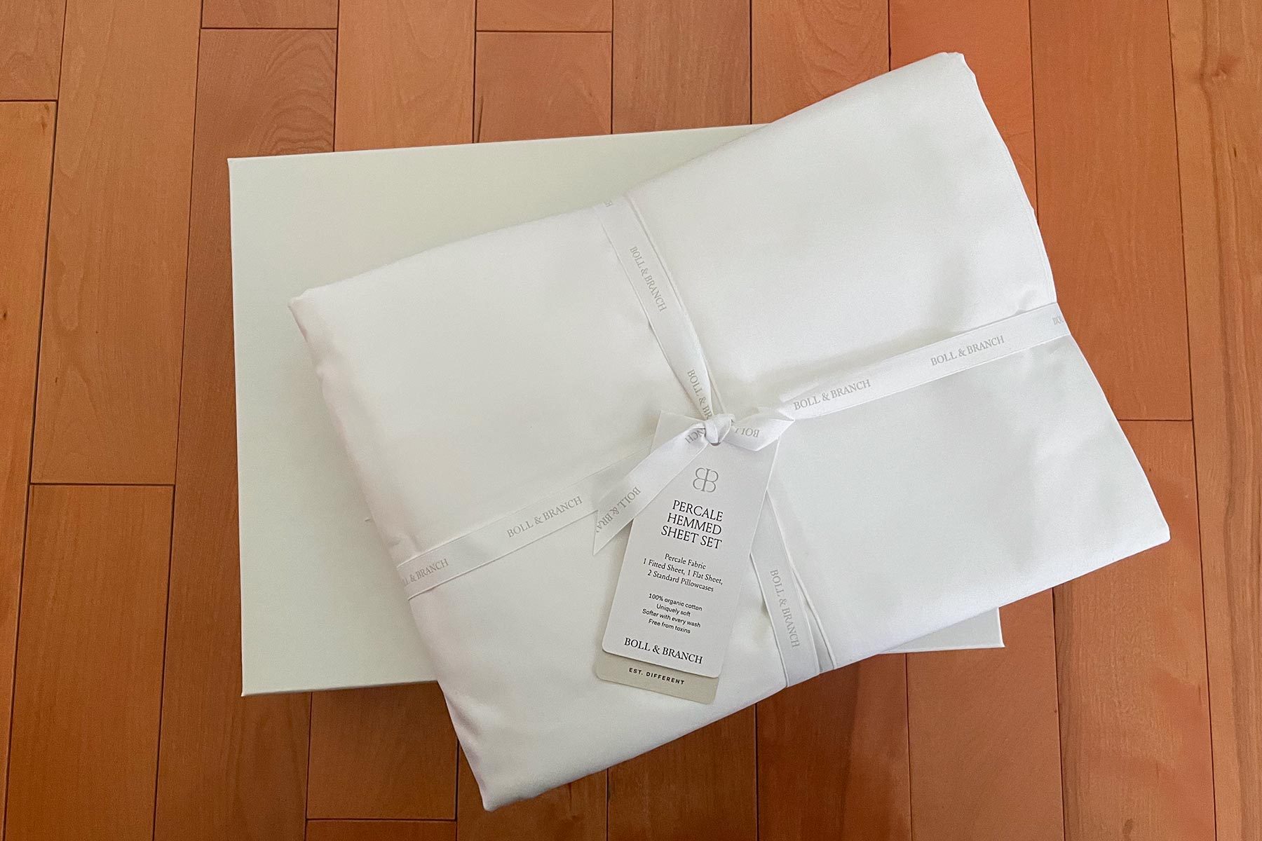 Sheet set wrapped in ribbon, lying on a wooden floor. Tag reads "Percale Hemmed Sheet Set, Boll & Branch, 100% organic cotton."