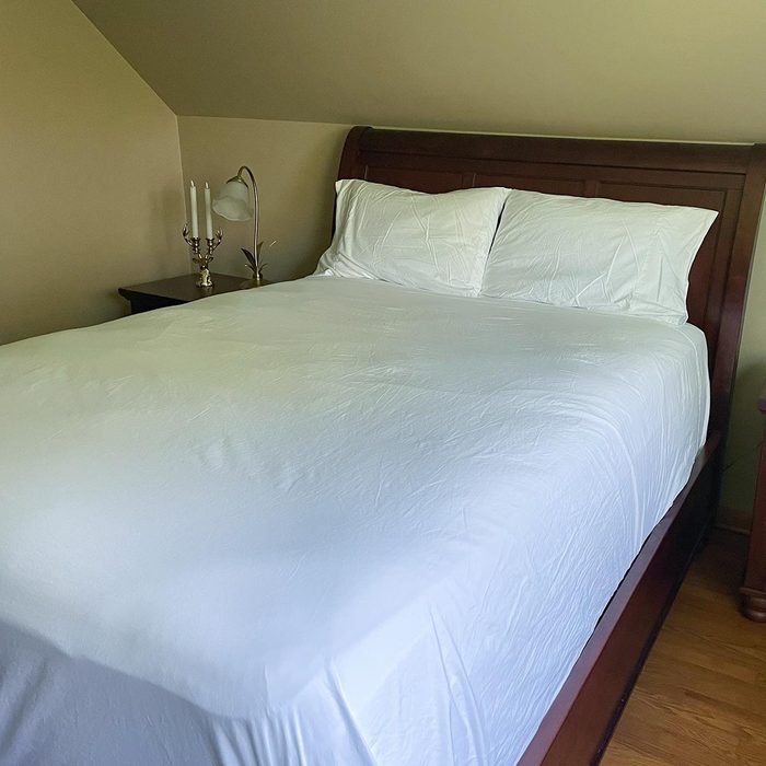 Bed neatly made with white sheets, positioned in a corner bedroom beside a nightstand with a lamp and decorative candleholder.